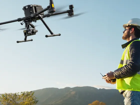 Un homme portant un casque blanc, un gilet jaune de sécurité, contrôle un drone dans un paysage avec des montagnes et un arbre.
