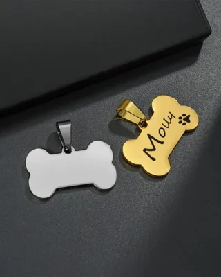 Deux pendentifs en forme d'os, un en or avec le nom 'Mowy' et une empreinte de patte, l'autre en argent, posés sur une surface noire avec une boîte noire en arrière-plan.