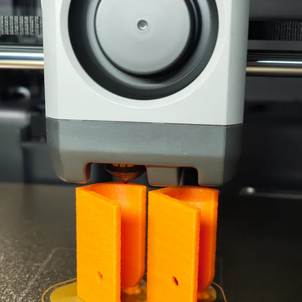 Une imprimante 3D déposé deux pièces orange en forme de rectangle pendant le processus d'impression.