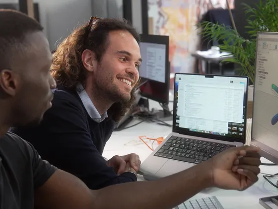 Deux hommes discutent devant des ordinateurs dans un bureau moderne, avec un logiciel de modélisation 3D affiché sur un écran.