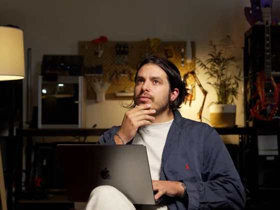 Un homme assis à un bureau avec un ordinateur portable, en train de réfléchir, dans une pièce avec des étagères contenant des bobines de fil, une lampe de lecture, des plantes et des outils.
