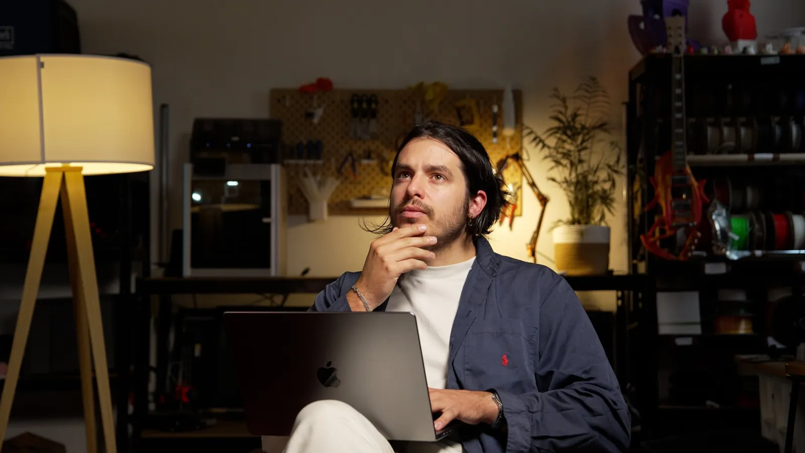 Un homme avec des cheveux longs assis devant un ordinateur portable, dans un atelier ou un studio, réfléchissant avec la main sur le menton.