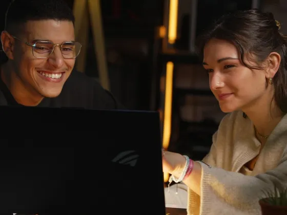 Deux jeunes adultes, un homme avec des lunettes et une femme avec des boucles d'oreilles dorées, regardant un ordinateur portable avec sourires, dans un environnement intérieur chaleureux.