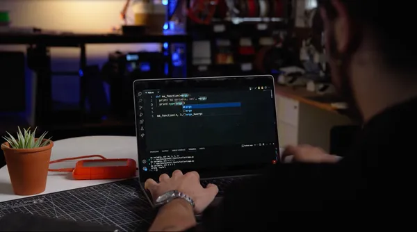 Person travaillant sur un ordinateur portable avec un environnement de bureau sombre et des étagères en arrière-plan, en train de coder en Python.