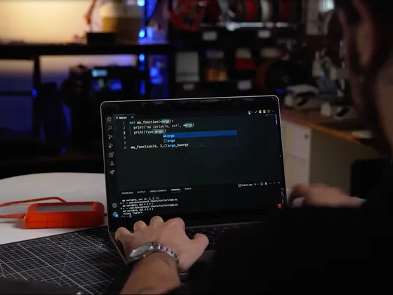 Une personne travaille sur un ordinateur portable avec du code Python affiché à l'écran, sur une table blanche avec une plante en pot, dans un environnement de bureau ou atelier.