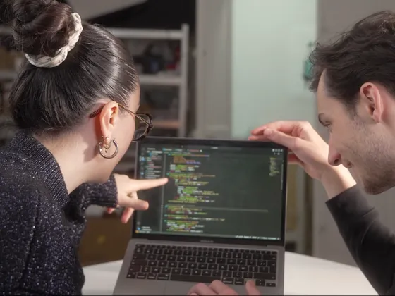 Deux personnes, une femme avec des cheveux relevés, portent des lunettes et des boucles d'oreilles, et un homme, regardent un écran d'ordinateur portable affichant du code. Elles semblent collaborer sur un projet de programmation.