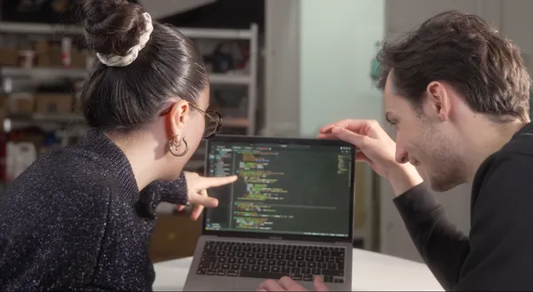 Deux personnes regardent un écran d'ordinateur avec du code informatique coloré, en discutant dans un environnement de bureau ou atelier.