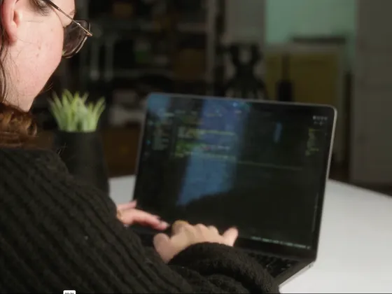 Une personne avec des cheveux bruns et porte des lunettes, utilisant un ordinateur portable dans un environnement intérieur, probablement un bureau ou un espace de travail.