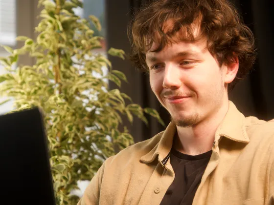 Jeune homme avec des cheveux bouclés, portant une chemise beige, souriant en regardant un ordinateur portable dans un intérieur lumineux avec une plante verte en arrière-plan.