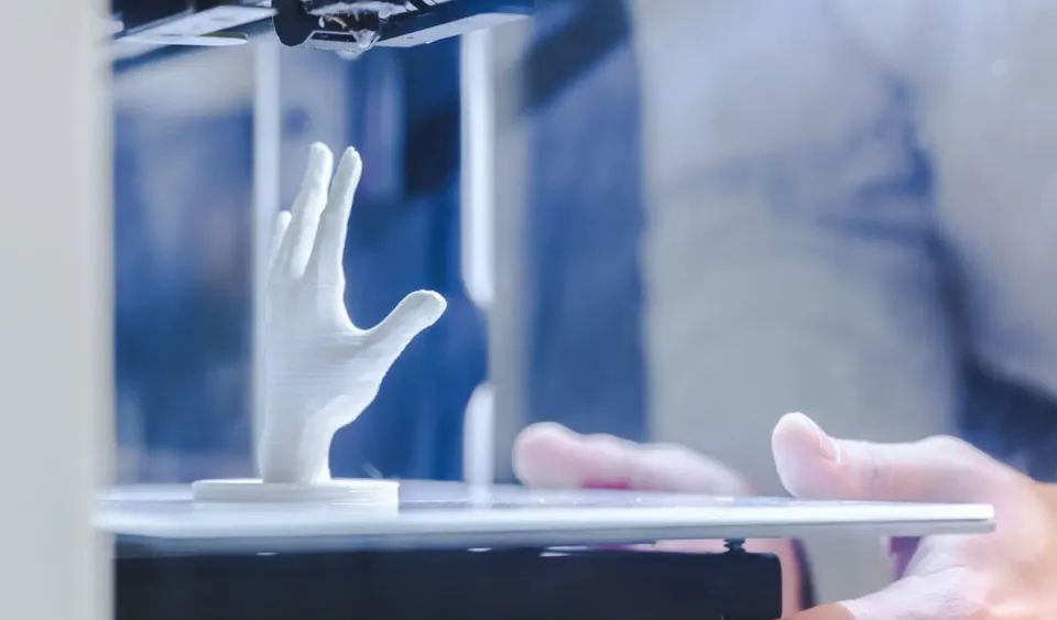Une main en gants blancs approche une main en plastique d'une imprimante 3D.