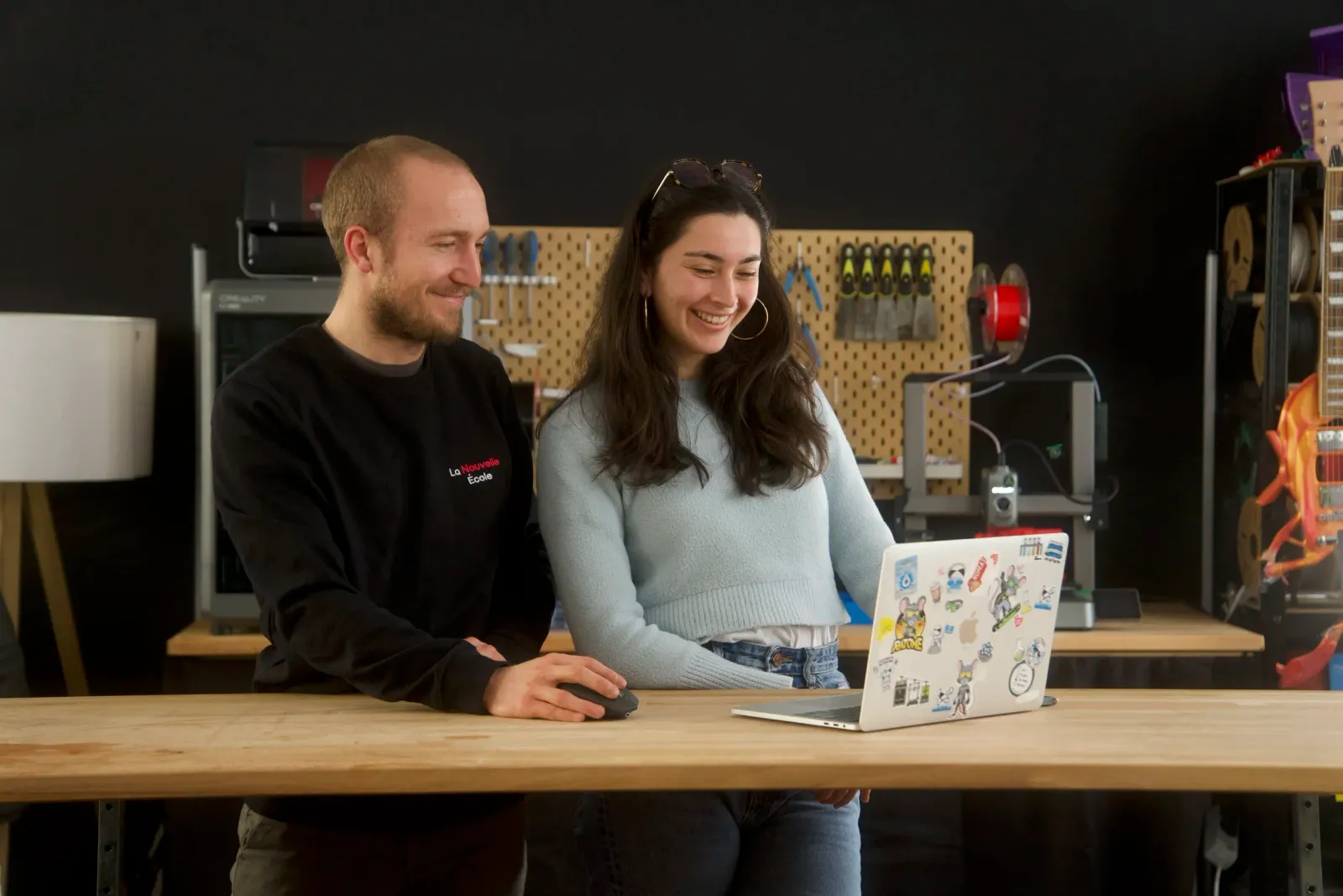 Deux personnes souriantes regardant un ordinateur portable recouvert de stickers dans un atelier.