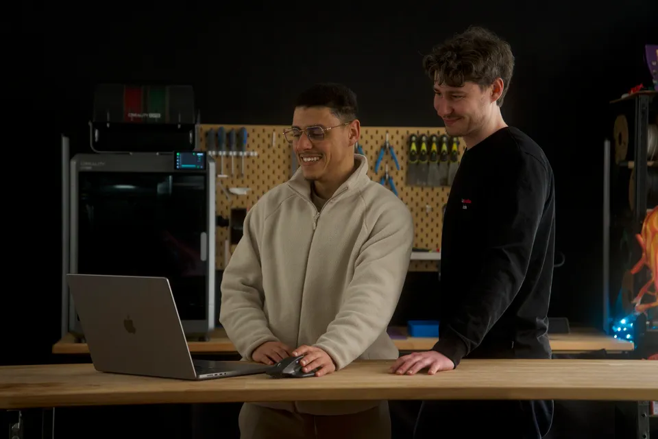 Deux jeunes hommes souriants se regardent devant un ordinateur portable sur un plan de travail en bois dans un atelier ou un espace de travail technologique.