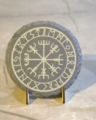 Pierre ronde gravée de symboles runiques et de signes anciens, exposée sur un support en métal, posé sur une surface en pierre ou en céramique.