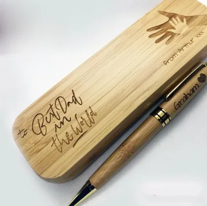 Un stylo en bois avec gravure, posé sur un étui en bois gravé avec une main et une inscription. La gravure sur l'étui dit `to Beatrice my Thrilled` et la gravure sur le stylo dit `Graham`.