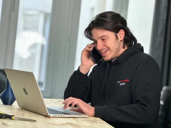 Jeune homme avec cheveux longs, portant un sweat noir avec le logo 'La Nouvelle École', parlant au téléphone portable, assis à une table avec un ordinateur portable.