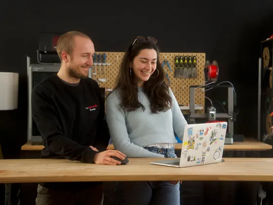 Deux personnes, un homme et une femme, regardent un ordinateur portable et sourient dans un atelier technologique.