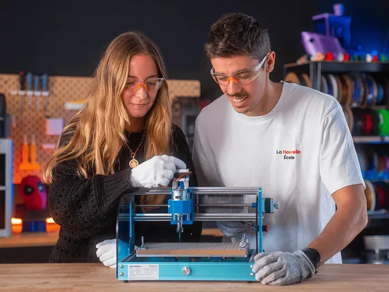 Deux personnes portant des lunettes de protection et des gants travaillent sur une machine de fabrication additive dans un atelier. On voit des bobines de fil en arrière-plan.