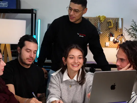 Groupe de jeunes adultes en discussion autour d'un ordinateur portable dans un atelier ou école technologique.