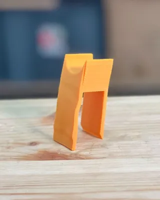 Une petite pièce en plastique orange en forme de pince ou de support, posée sur une surface en bois.