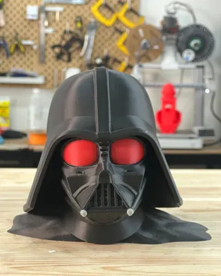 Figurine en forme de casque de Darth Vader de Star Wars avec des yeux rouges, placée sur une table en bois, avec un atelier de bricolage en arrière-plan.