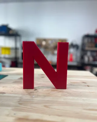 Lettre en plastique rouge en forme de N posée sur un plan de travail en bois, avec un fond flou d'étagères industrielles et d'objets divers.