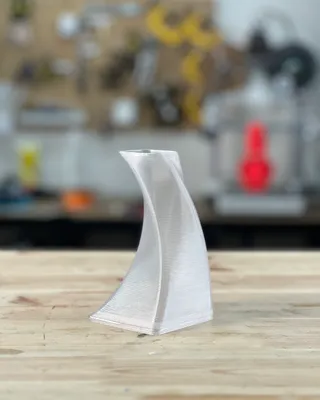 Une sculpture en plastique transparent et ondulée sur une table en bois, avec un fond flou comprenant des outils et des objets de bureau.