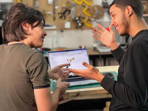 Deux hommes discutent dans un atelier avec un ordinateur portable, en montrant un croquis ou un diagramme. Le mur derrière eux est équipé d'établi avec des outils, notamment des scies circulaires et autres outils électriques.