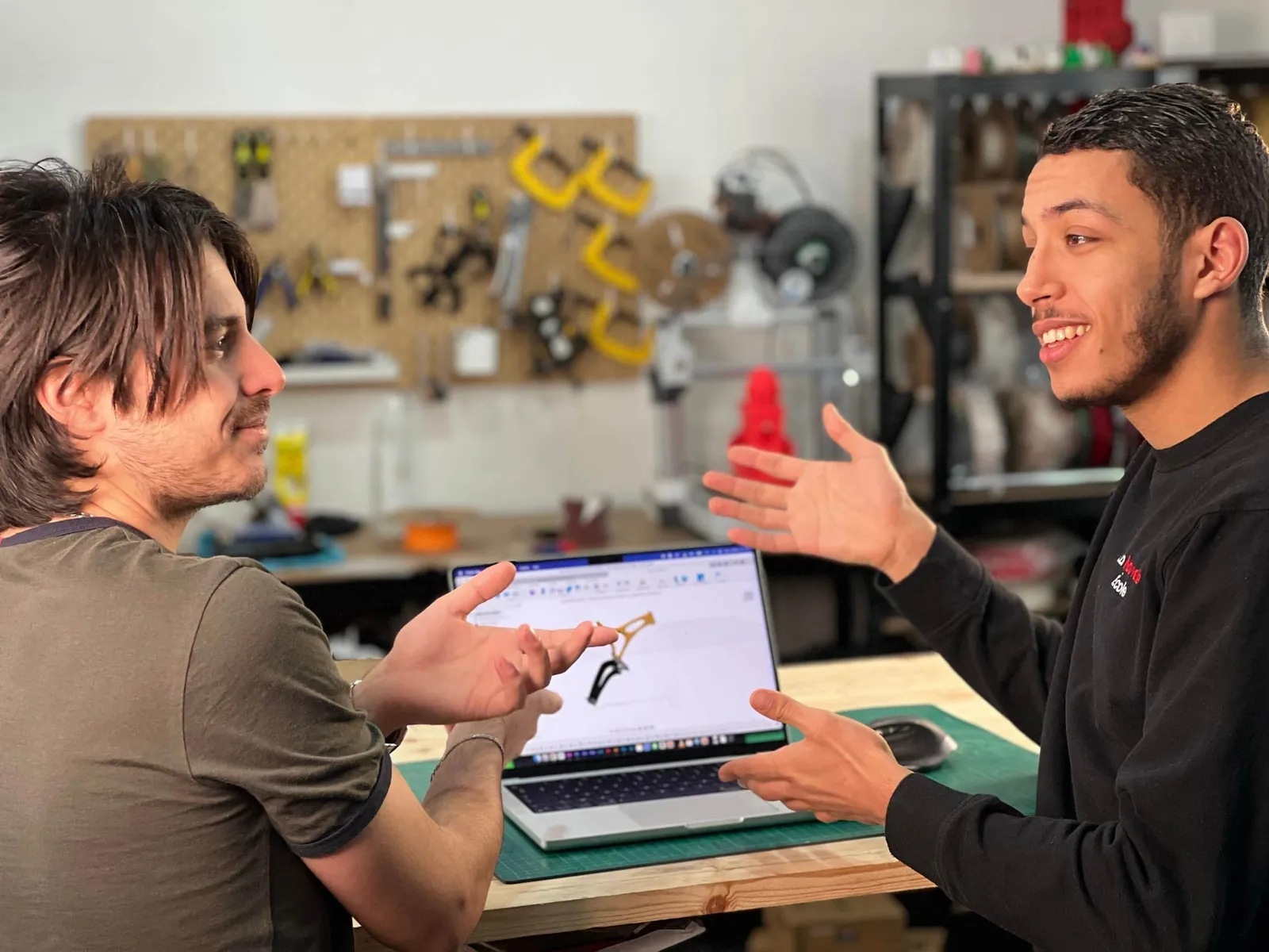 Deux hommes discutent dans un atelier avec des outils suspendus au mur en arrière-plan, l'un montre un modèle de pièce détachée sur un ordinateur portable.