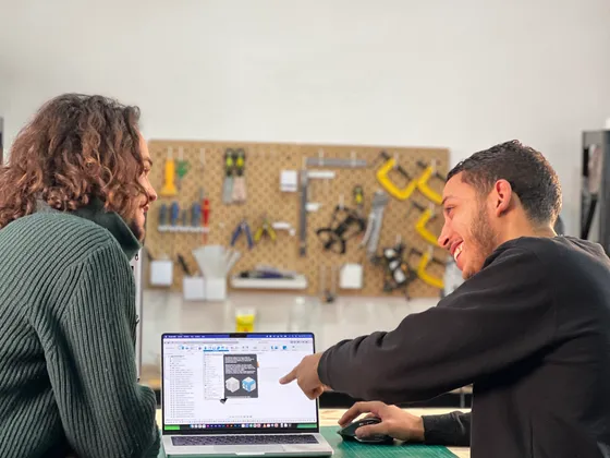 Deux hommes discutent dans un atelier ou un espace de travail, un d'eux pointe un ordinateur portable avec des images de cubes 3D à l'écran, arrière-plan avec un panneau d'outils et matériel de bricolage.