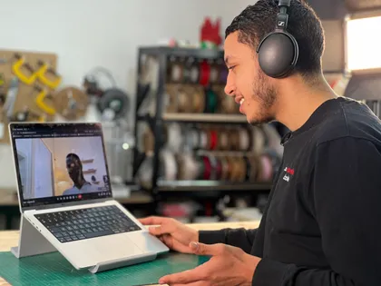 Un jeune homme portant un casque tourne un appareil photo vers un ordinateur portable devant lui, dans un atelier avec des rouleaux de fil ou de câbles en arrière-plan.