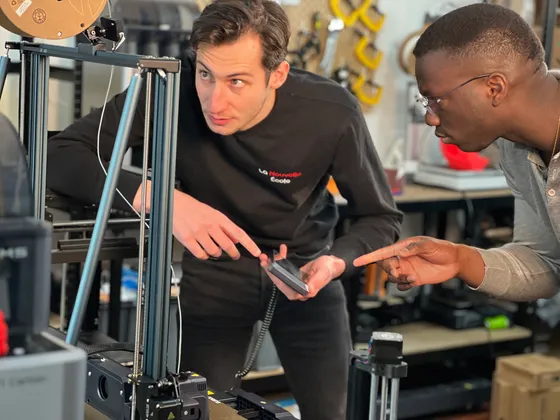 Deux hommes discutent devant une imprimante 3D dans un atelier technique, l'un pointant avec un téléphone portable, l'autre observant attentivement.