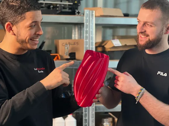 Deux jeunes hommes souriants se montrent une pièce rouge en forme de spirale dans un atelier. Sur l'étagère derrière eux, on voit des boîtes étiquetées avec des mots comme 'Câbles', 'Acétone' et 'Peinture'.
