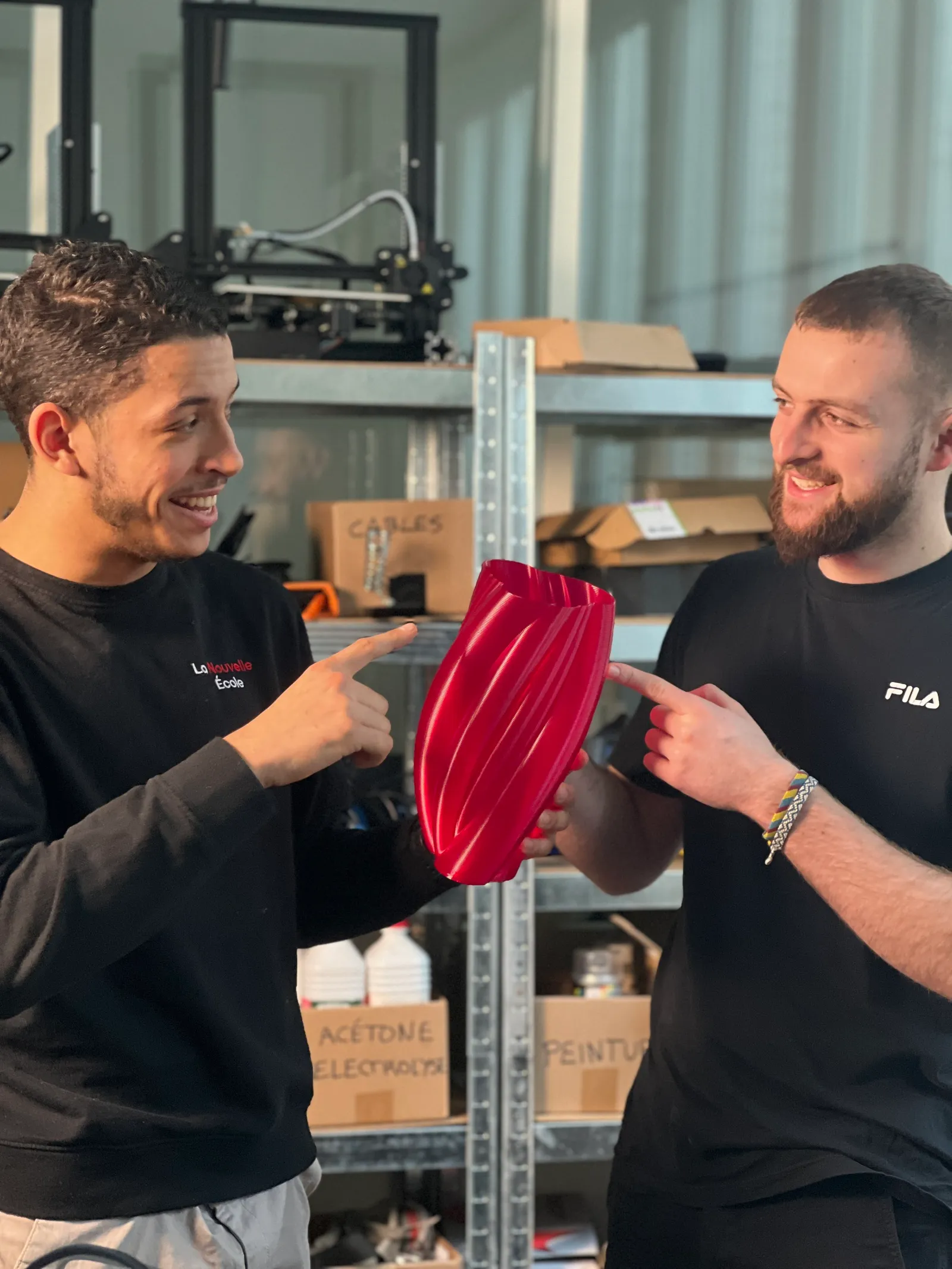 Deux jeunes hommes souriants se montrent une pièce rouge imprimée en 3D dans un atelier