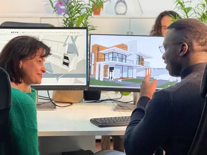 Deux personnes discutent devant des écrans d'ordinateur affichant des plans architecturaux, dans un environnement de bureau avec des plantes en arrière-plan.