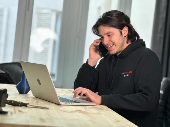 Jeune homme souriant, parlant au téléphone, assis à une table avec un ordinateur portable.