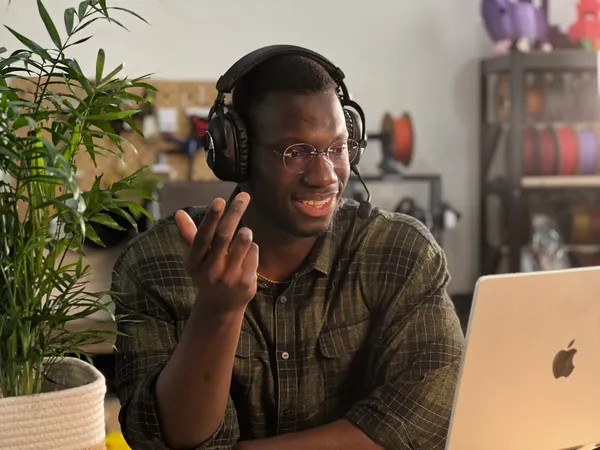 Homme souriant portant un casque avec microphone, en train de parler ou d'écouter, assis devant un ordinateur portable Apple dans un environnement intérieur avec une plante verte à gauche et une étagère en arrière-plan.