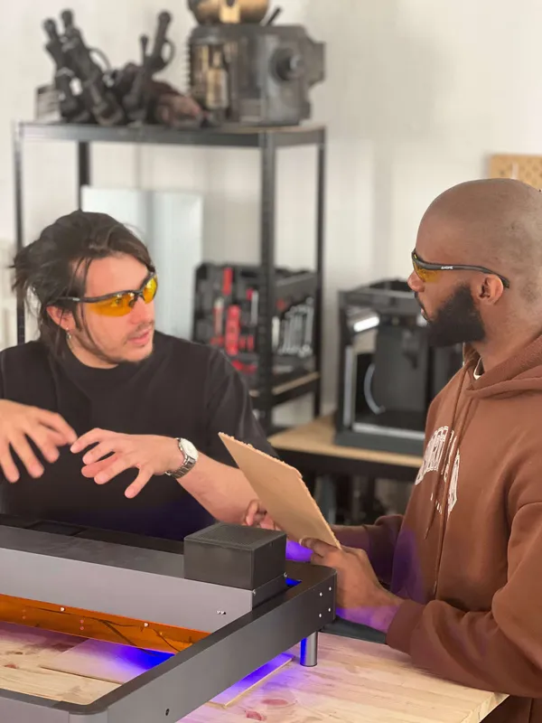 Deux hommes discutant dans un atelier, portant des lunettes de protection jaunes, avec des outils et des machines en arrière-plan.