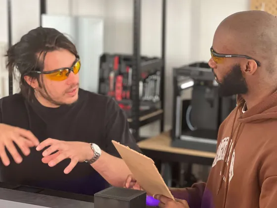 Deux hommes discutent dans un atelier, portant des lunettes de sécurité jaunes, avec une machine en science et des outils en arrière-plan.