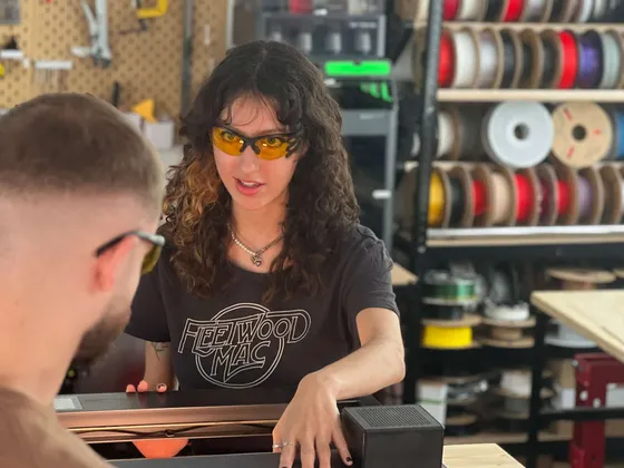 Une femme avec des cheveux bouclés, portant des lunettes de protection orange, explique quelque chose à un homme à côté d'une imprimante 3D dans un atelier. Elle porte un t-shirt noir 'Fleetwood Mac' et se trouve devant des étagères remplies de rubans ou filaments pour imprimantes 3D.