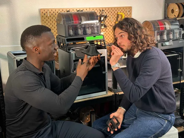 Deux hommes discutent dans un atelier technologique, l'un montre un objet en plastique à l'autre, entourés d'équipements électroniques et outils.