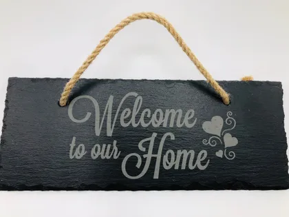 Plaque en ardoise noire avec l'inscription "Welcome to our Home" en gris, ornée de cœurs, suspendue par une corde en jute.