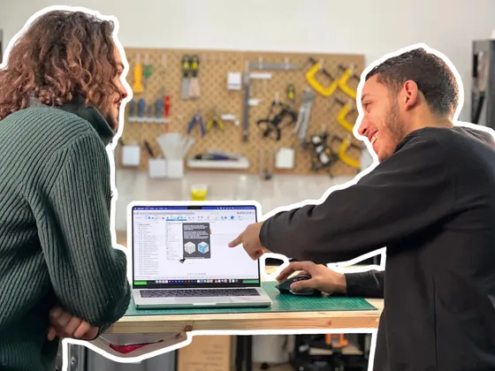 Deux hommes discutent devant un ordinateur portable dans un atelier, en train de montrer quelque chose à l'écran, avec un mur de supports à outils en arrière-plan.