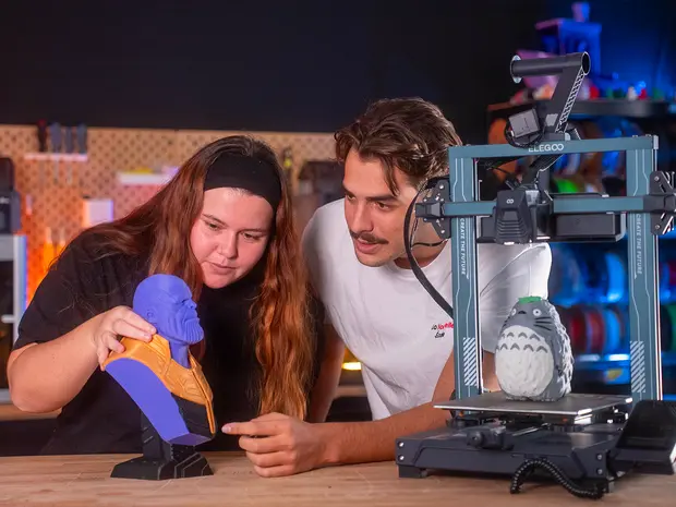 Deux personnes travaillent sur une imprimante 3D dans un atelier, une femme tient une figurine de Totoro, un personnage du film d'animation japonais, tandis qu'un homme regarde.'}
