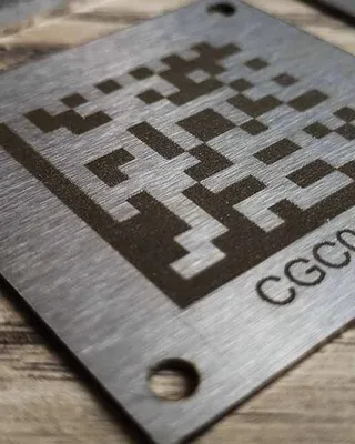 Plaque en métal avec un motif QR code gravé, posée sur une surface en bois.