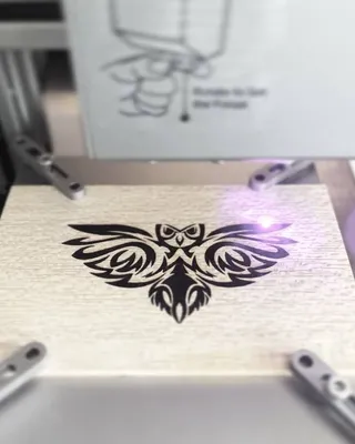 Une machine laser gravant une image d'une chouette stylisée sur un panneau en bois clair.