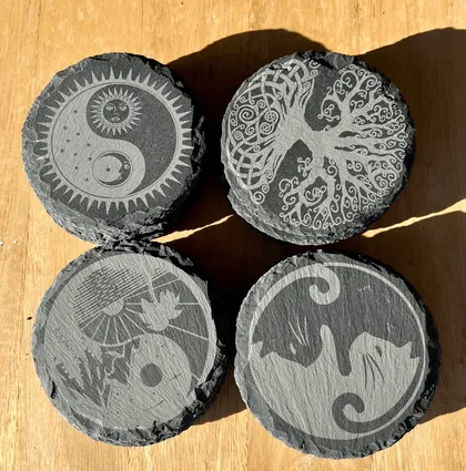 Quatre pierres plates gravées avec des symboles mystiques, dont un symbole yin-yang, un arbre de vie, un soleil et lune, et un dragon, disposées sur une surface en bois.