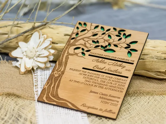 Faire-part de mariage en bois avec découpe d'arbre, posé sur un tissu en toile de jute, avec décoration florale blanche et branche sèche en arrière-plan.