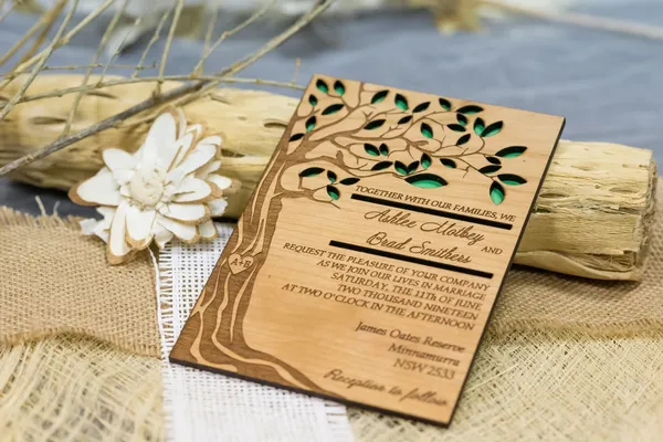 Invitation de mariage en bois gravé avec arbre et feuilles, placée sur un tissu en toile avec une fleur en bois et un morceau de bois flotté en arrière-plan.