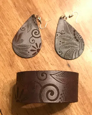Un bracelet, une paire de boucles d'oreilles en cuir avec motifs floraux et un bracelet large assorti, tous sur une surface en bois.