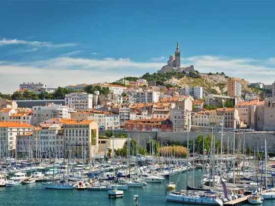 Port de plaisance avec de nombreux yachts et voiliers, ville urbaine avec immeubles aux toits en tuiles rouges et collines en arrière-plan, avec la basilique Notre-Dame de la Garde au sommet.